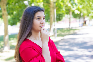 Pensive pretty young woman touching chin in park. Beautiful lady wearing red blouse and looking away with green trees in background. Contemplation and nature concept.