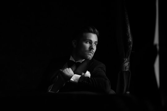 The fashionable bridegroom expects the bride near the window. Black and white portrait of the groom in a black suit