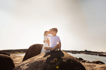A little boy sits on a stone and looks at the sea. His parents with a little sister stand behind and hug