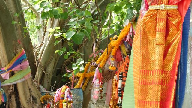 Colorful offering clothes on tree near altar. Bunch of bright traditional clothes as gift to Thai spirits hanging on tree branches near animist altar.