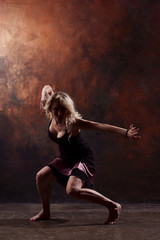 Photo of young dancing blonde woman with hands to sides in black dress on brown background