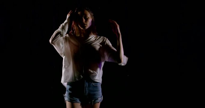 Close-up portrait of a blonde girl model who is in a dark Studio on a black background in heavy rain and posing. She's wearing a wet white shirt and short denim shorts.