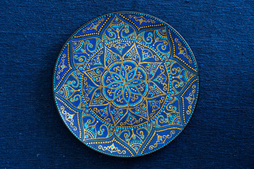 Decorative ceramic plate with blue and golden colors, painted plate on the background of blue fabric, closeup. Decorative porcelain plate painted with acrylic paints, handwork