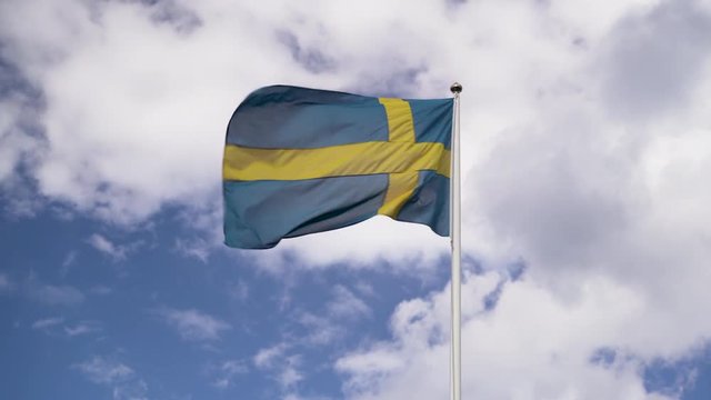 Swedish flag blowing in the wind against a blue sky and white clouds.