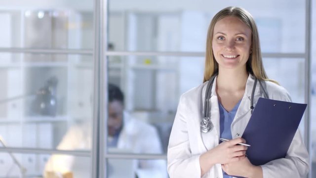 Waist-up portrait shot of 30-something Caucasian female physician, wearing blue scrubs and white lab coat, with stethoscope around neck and clipchart, standing in hospital doctors room and smiling