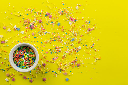 Cake topping prinkles on bright yellow background with little white bowl with various type of coloured sprinkles at bottom  and multi-colored cake topping sprinkles and star shapes sprayed around
