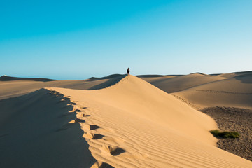 Fototapeta na wymiar evocative young confident woman walking on her own footprints path on the desert on top of dune with red dresson hot summer day with clear blue sky