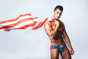 Portrait of handsome young man with stylish haircut in swinwear posing with American flag over gray...