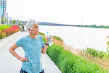 Portrait of fit and sporty mature woman drinking water. Shot of beautiful female runner standing outdoors holding water bottle. Fitness woman taking a break after running workout.