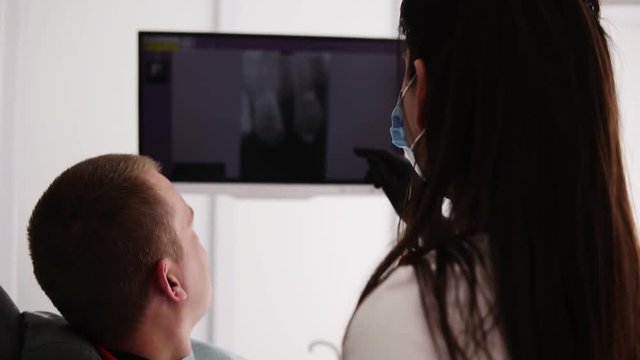 Caucasian female dentist explaining x-ray image on the screen to male patient. Shows the image of unhealthy tooth to a client. Footage from the back