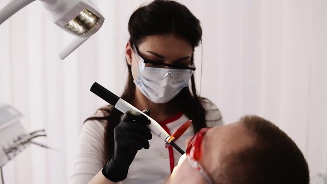 Female doctor and male patient in protective orange glasses in the modern dental ocabinet. Dentist is doing polymerisation of fillings with a orange protective screen. Overview footage, slow motion