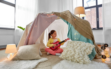 childhood and hygge concept - happy little girl playing guitar in kids tent or teepee at home