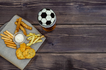 Diverse snacks and beer with a soccer ball on a beer foam on dark wooden background. Top view, Empty space for text
