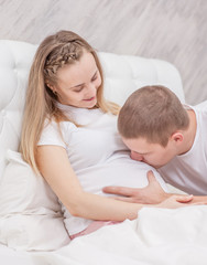 Young man kisses to the belly of his pregnant wife on the bed