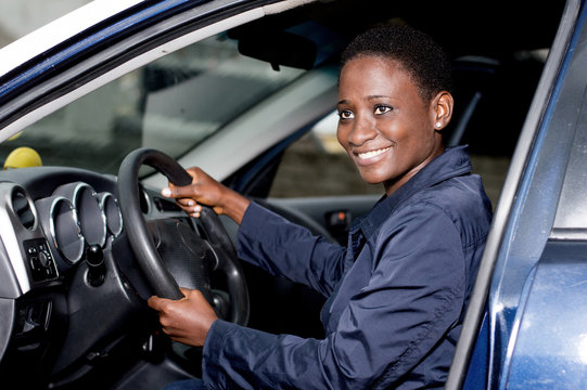 Young woman sitting in a car, hands on the steering wheel smiling at the camera.