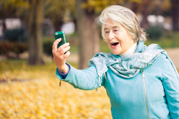 View of a smilling and espressive pretty senior woman looking at his phone on yellow leaves in front of a trees with autumnal colors.