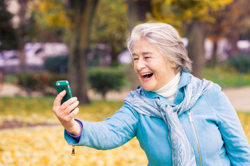 View of a smilling and espressive pretty senior woman looking at his phone on yellow leaves in front of a trees with autumnal colors.