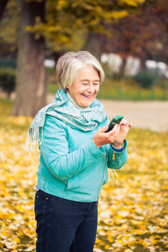 View of a smilling pretty senior woman with his phone on yellow leaves in front of a trees with autumnal colors.