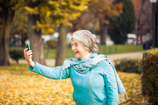 View of a smilling pretty senior woman making a selfie with his phone on yellow leaves in front of a trees with autumnal colors.