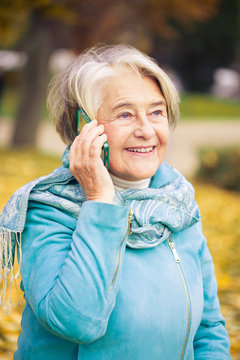 Sunny portrait of a smilling pretty senior woman calling with his phone in front of a tree with autumnal colors.