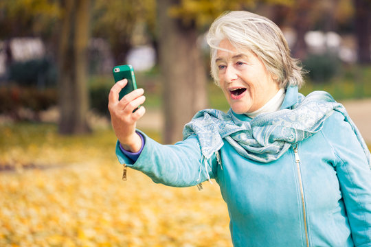 View of a smilling and espressive pretty senior woman looking at his phone on yellow leaves in front of a trees with autumnal colors.