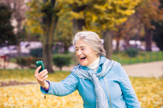 View of a smilling and espressive pretty senior woman looking at his phone on yellow leaves in front of a trees with autumnal colors.