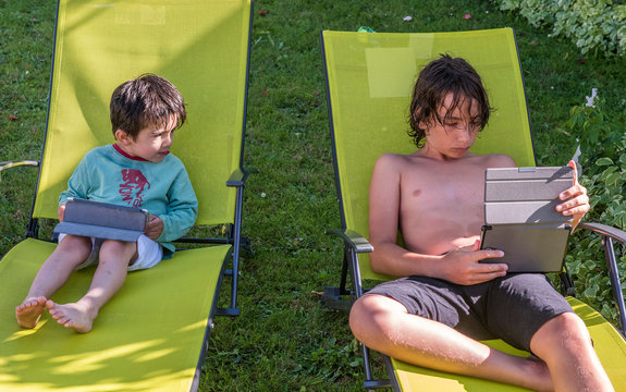 Two boys of four years old and twelve years old, sitting on deckchairs with their digital tablet