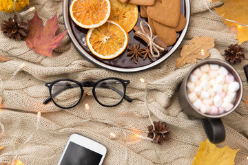 technology and season concept - smartphone, autumn leaves, hot chocolate, gingerbread cookies and glasses with garland lights on warm knitted blanket