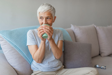 Happy lady relaxing at home with hot drink.  Senior woman holding coffee mug sitting on sofa and thinking.   Thoughtful lady sitting comfortably at home in autumn.