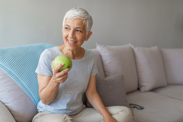 Stay healthy and wealthy. Close up of a smiling senior woman eating an apple at home. Age, healthy eating, food, diet and people concept - Happy smiling senior woman with green apple at home