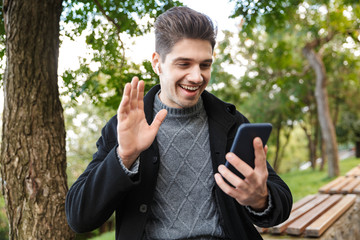 Handsome young man in casual clothing walking outdoors in green park using mobile phone listening music with earphones waving to friends talking.