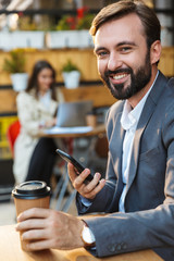 Portrait of smiling happy man drinking takeaway coffee and using mobile phone while sitting in cafe