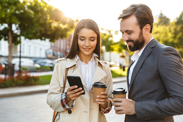 Photo of beautiful office workers man and woman holding takeaway coffee while using mobile phone on city street