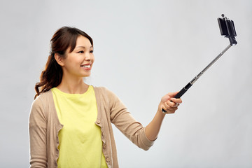 technology and people concept - happy asian woman taking picture by smartphone on selfie stick over grey background