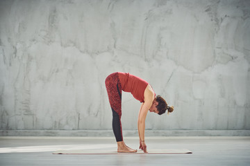 Fototapeta Side view of beautiful Caucasian brunette in red sports wear standing barefoot on the mat in Standing Forward Bend yoga posture. obraz