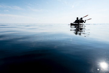 Silhouettes of two tourists going in a kayak on the smooth water of the lake, against the sky. Activities and sports.