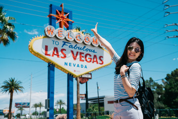 Happy young girl backpacker in sunglasses showing peace gesture and looking at camera while standing by famous billboard in nevada. smiling lady traveler taking picture with las vegas sign on summer