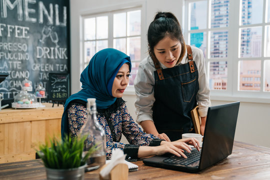 Young Successful Malay Woman Employee Small Business Owner Sitting In Morning Coffee Shop. Asian Girl Waitress Staff Talking With Friend Client Together Doing Project Giving Advice On Laptop Pc.