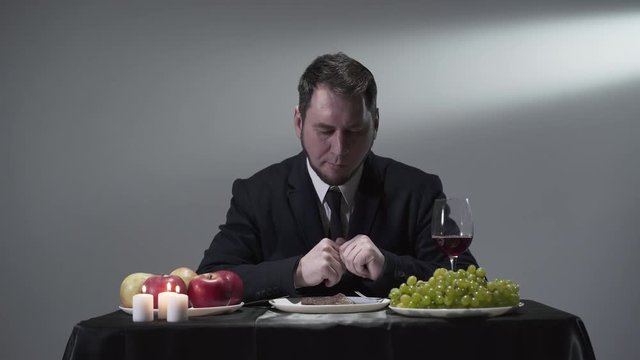 Rich businessman in a suit eatting or tasting a piece of meat next to apples, grapes and a glass of red wine.