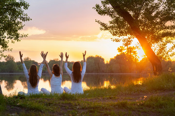 beautiful young women doing yoga in white clothes arms raised to the sky in prayer, at sunset by a...