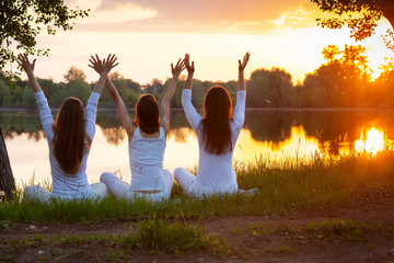 Women doing yoga in white clothes arms raised to the sky in prayer, at sunset by a large lake...