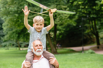 Little boy with airplane model and grandfather raising hands over green park on background enjoying life and nature. Portrait of happy grandfather giving grandson piggyback ride on his shoulders.