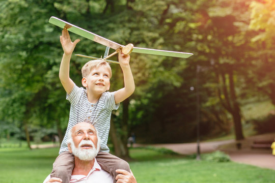 Child boy with airplane model and grandfather raising hands over sunset enjoying life, nature. Happy grandfather giving grandson piggyback ride on his shoulders and looking up. Sun glare effect.
