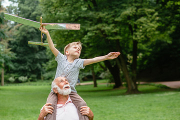 Little boy with airplane model and grandfather raising hands over green park on background enjoying life and nature. Portrait of happy grandfather giving grandson piggyback ride on his shoulders.