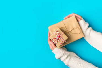 Young Caucasian woman in knitted white sweater holds in hand stack of gift boxes wrapped in brown Kraft paper on blue background. Christmas New Year presents concept. Eco friendly materials