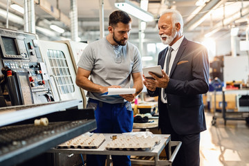 Smiling mature engineer and worker using touchpad in factory plant.