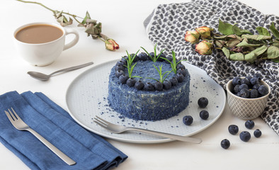Table with Blue Blueberry Velvet cake. Cup of coffee with milk, small bowl with blueberries, scarf and dry flowers, roses.