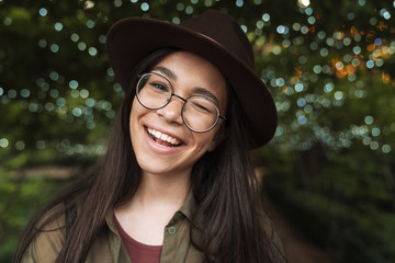 Photo closeup of young woman wearing hat and eyeglasses smiling with open mouth and winking while walking