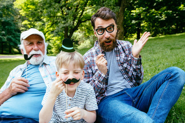 Happy funny family son and dad, granddad with fake mustache, hat, eyeglasses on holiday outdoor in...