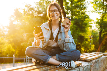 Happy cute young student girl wearing eyeglasses sitting outdoors in nature park using mobile phone drinking coffee.